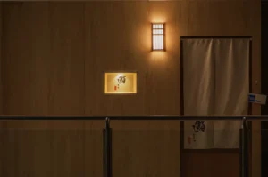 Dimly lit wooden hallway with a small illuminated sign featuring Japanese characters. A traditional curtain covers a doorway, creating a serene ambiance.