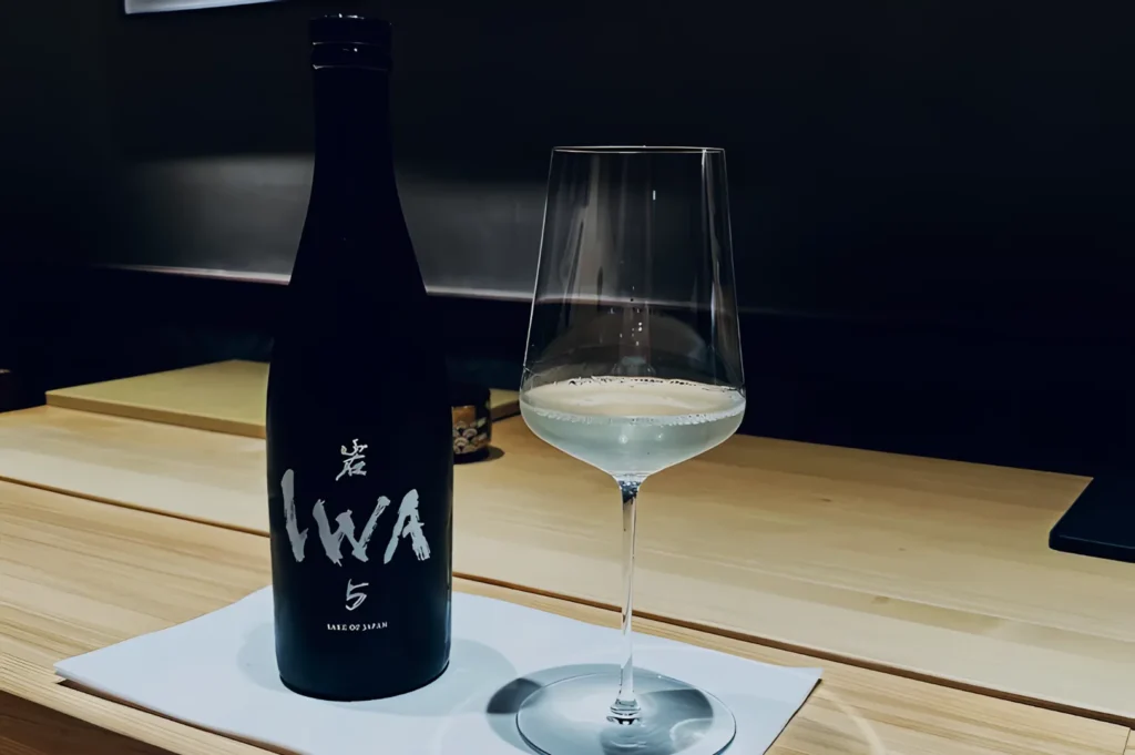 Black sake bottle labeled “IWA” and a half-filled wine glass on a light wooden counter, conveying elegance and sophistication in a dimly lit setting.