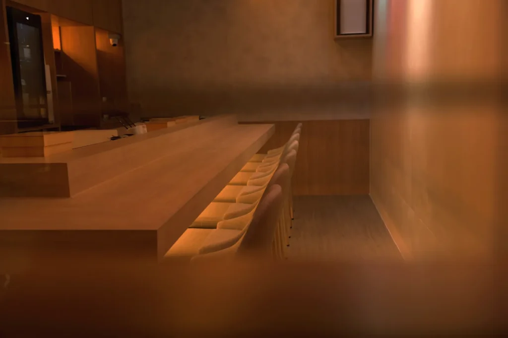 Dimly lit modern restaurant interior with a long wooden bar and padded stools. Warm, muted tones create a cozy, intimate atmosphere.