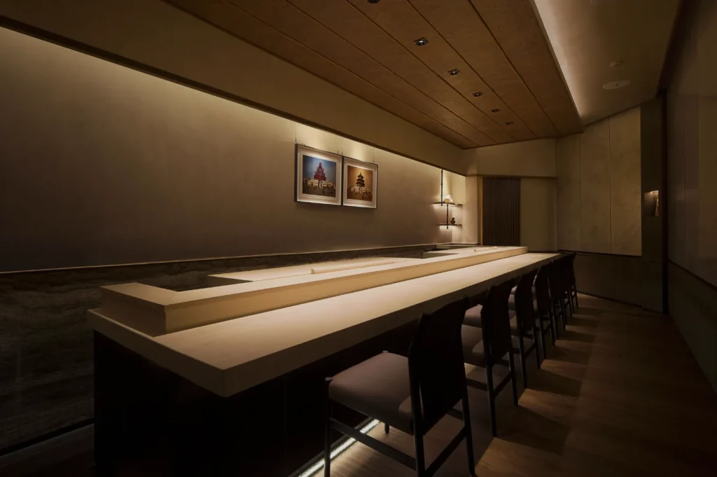 Elegant, minimalist sushi bar with a long light-wood counter and dark chairs. Dim lighting creates a serene ambiance; framed art on wall.