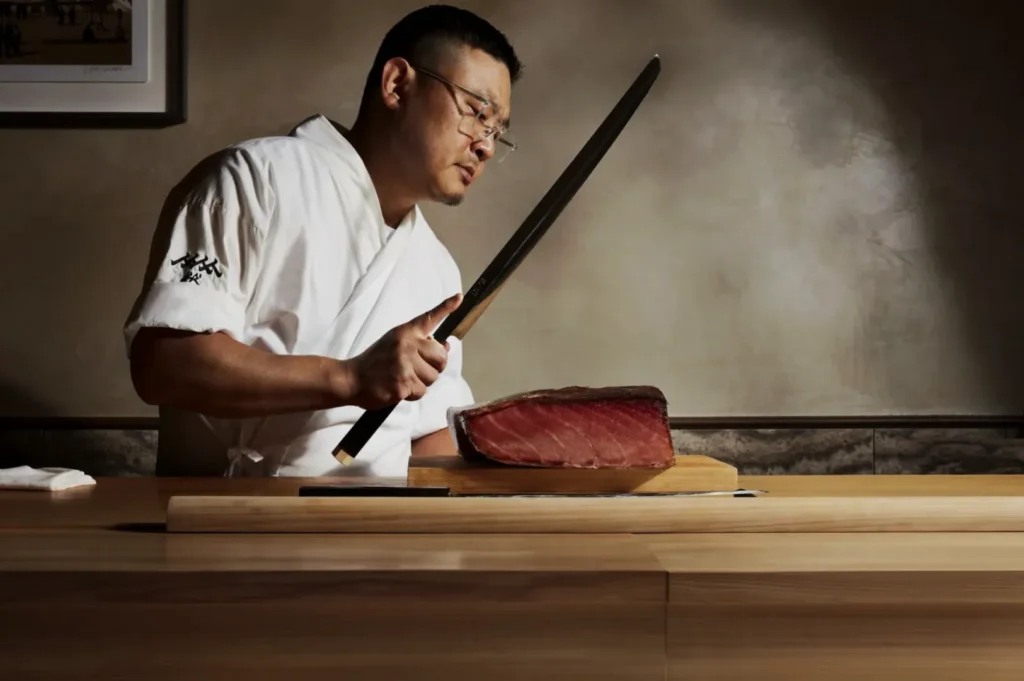 A sushi chef in a white uniform expertly holds a long knife, preparing to slice a thick piece of raw tuna on a wooden counter, creating a focused and professional atmosphere.