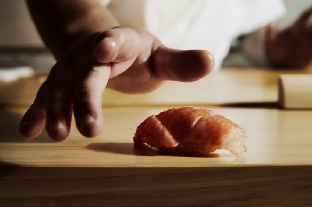 A sushi chef's hand poised over a piece of tuna nigiri on a wooden counter, capturing focus and precision in a traditional Japanese setting.