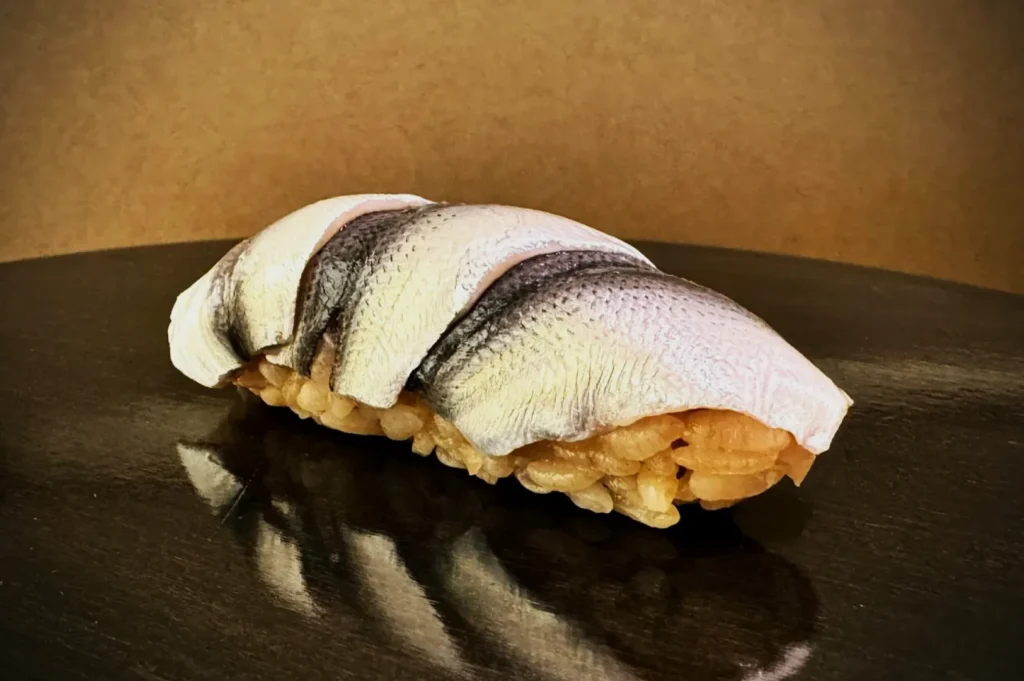 A piece of sushi is placed on a reflective black surface. The sushi features a slice of silver, textured fish atop seasoned rice, creating a fresh and elegant presentation.