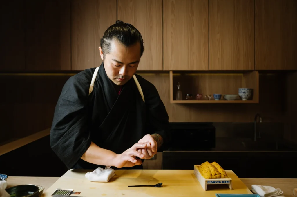 Chef in traditional black attire prepares sushi in a modern kitchen. With focused expression, he shapes rice near a box of sea urchin. Calm ambiance.