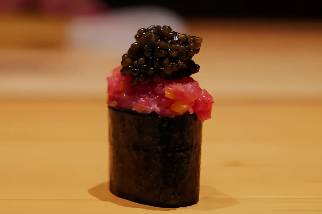 A piece of gourmet sushi on a wooden surface, featuring vibrant pink minced fish topped with black caviar, wrapped in nori, conveying elegance.