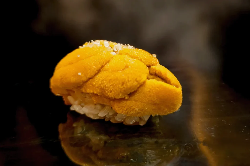 A piece of uni sushi sits on glossy black surface, featuring bright orange sea urchin atop a bed of white rice, dusted with salt crystals.