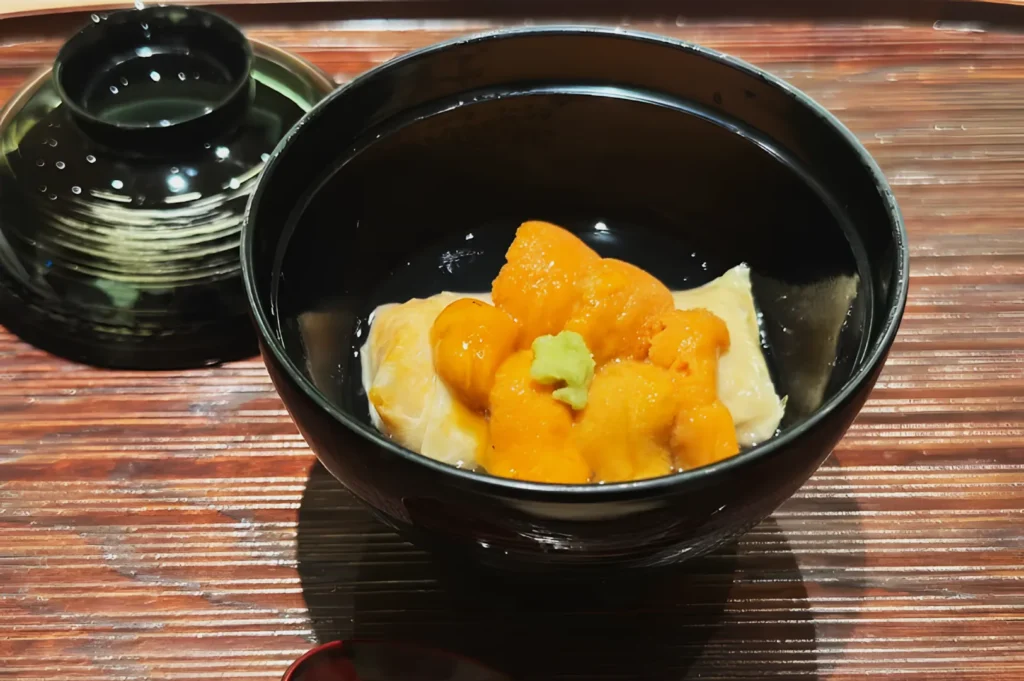 A black bowl with a wooden lid holds a Japanese dish, featuring vibrant orange sea urchin (uni) atop tofu, garnished with wasabi, on a wooden table.