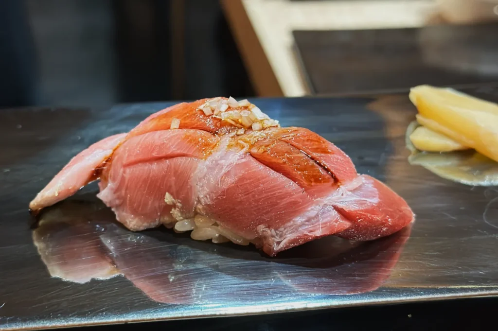 Close-up of fresh tuna nigiri sushi on a reflective plate, topped with finely chopped onions. A side of pickled ginger is visible, adding a gourmet touch.