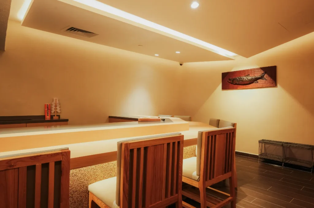 Minimalist sushi bar with wooden chairs facing a sleek counter under soft lighting. A fish painting adorns the cream-colored wall, creating a tranquil ambiance.