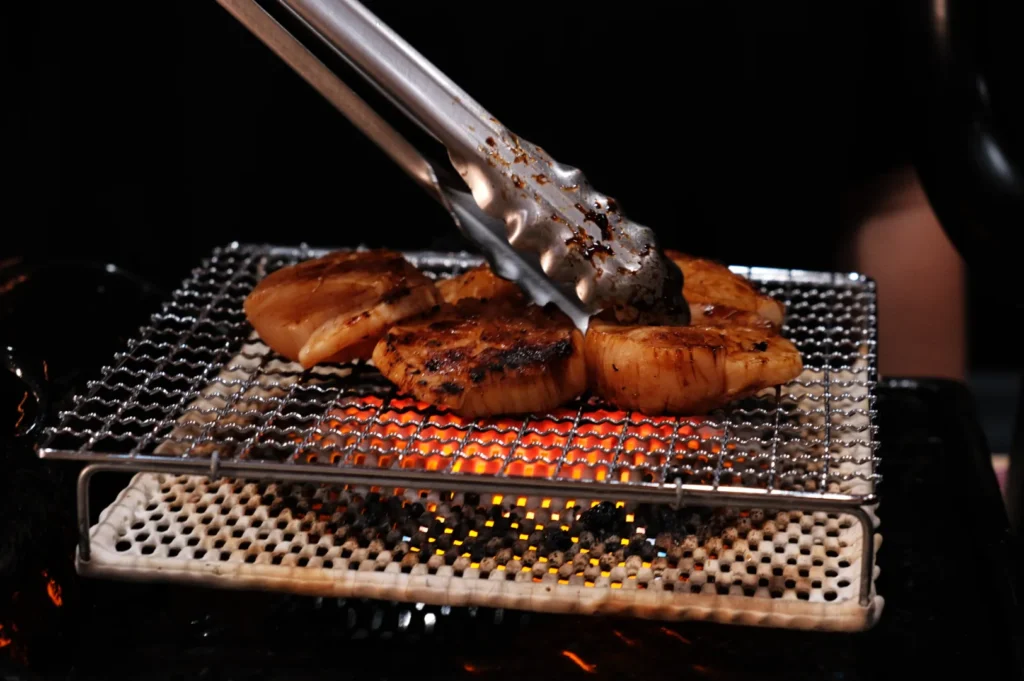 Grilled scallops being cooked on a mesh grill with glowing charcoal underneath. Tongs are used to flip them, capturing a warm and appetizing scene.