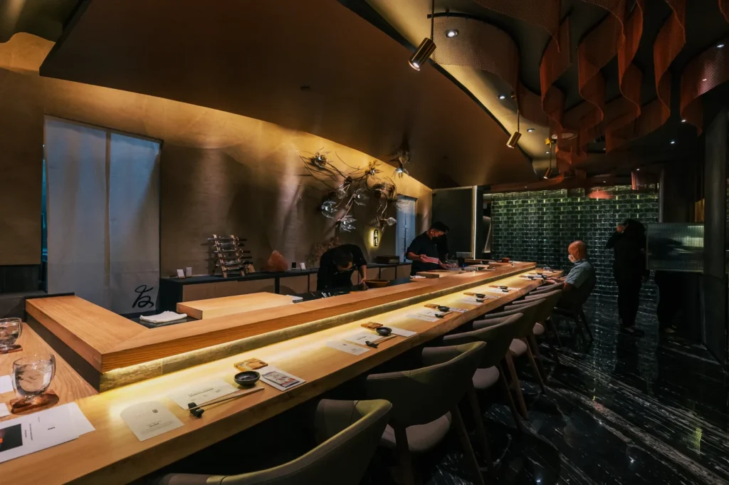 Elegant sushi bar with a long wooden counter, dim lighting, and modern decor. Two chefs prepare food while guests sit and enjoy the ambiance.