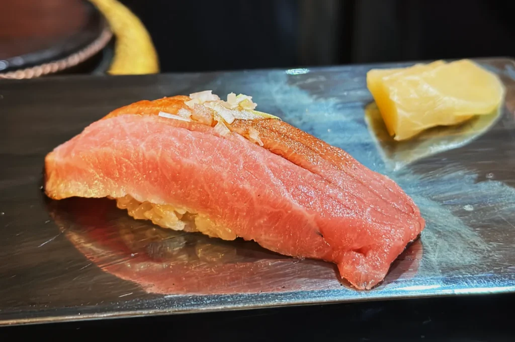 A piece of sushi with pink tuna on rice is garnished with finely chopped onions, served on a rectangular steel plate with a slice of yellow pickled ginger.
