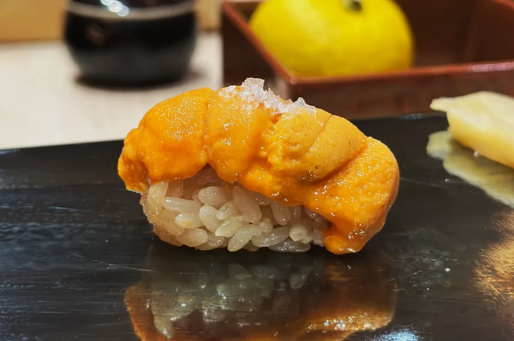 A piece of uni sushi sits on a glossy black plate. The vibrant orange uni contrasts with a mound of white rice. A lemon and soy sauce are blurred in the background.