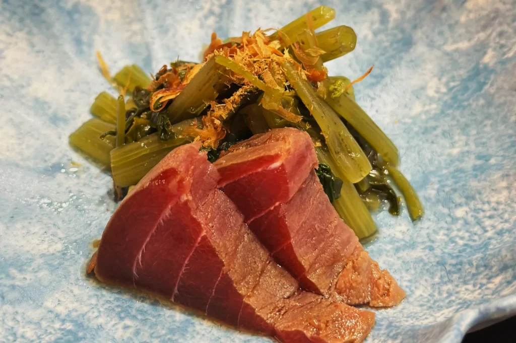 Sliced seared tuna arranged on a light blue ceramic plate, garnished with sautéed green vegetables and bonito flakes, conveying a fresh and elegant presentation.