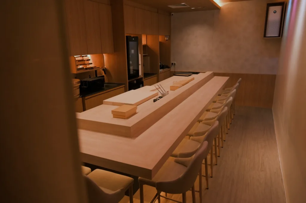 Minimalist sushi bar interior with a long wooden counter, light wood surfaces, and beige chairs. Warm lighting creates a cozy, serene atmosphere.