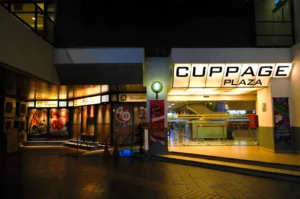 Entrance to Cuppage Plaza at night, with bright signage. Adjacent is a warmly lit Japanese restaurant featuring vivid food images, creating an inviting atmosphere.