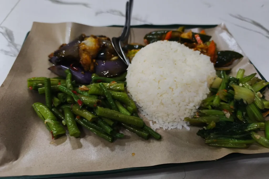 A serving of white rice is centered on a paper-lined plate, surrounded by green beans, sautéed eggplant, and mixed vegetables, creating a vibrant, appetizing meal.