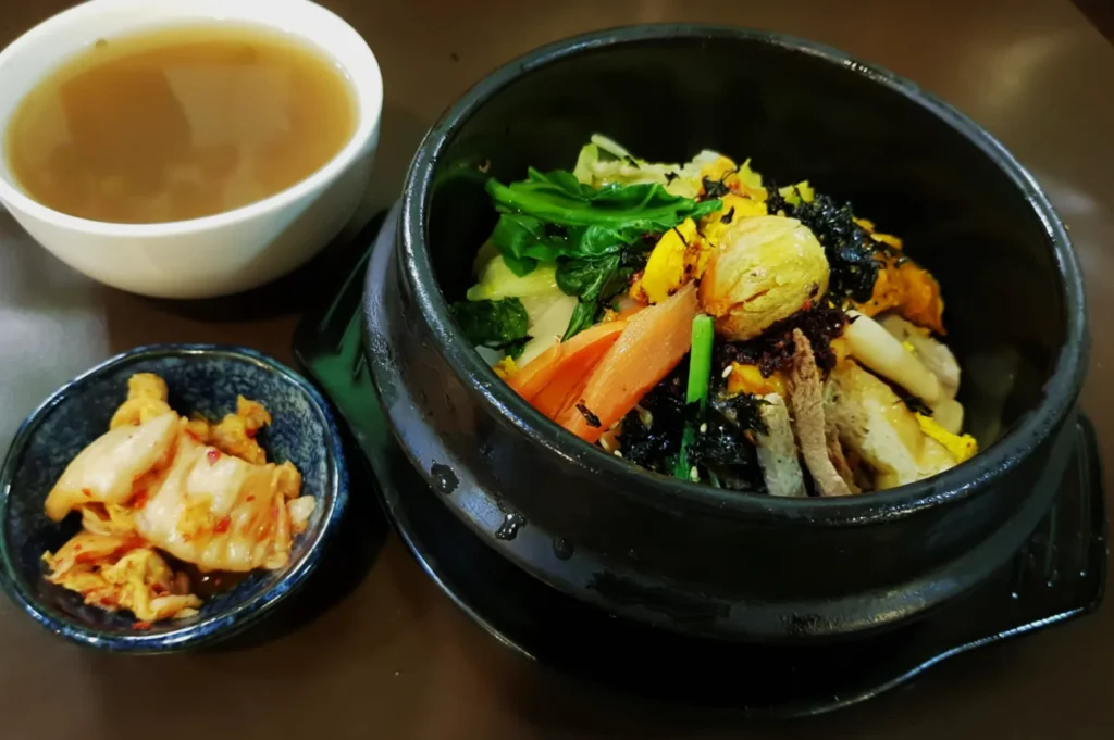 A rich Korean bibimbap in a black bowl, topped with vibrant vegetables and seaweed, served with kimchi and a bowl of broth on a brown table.