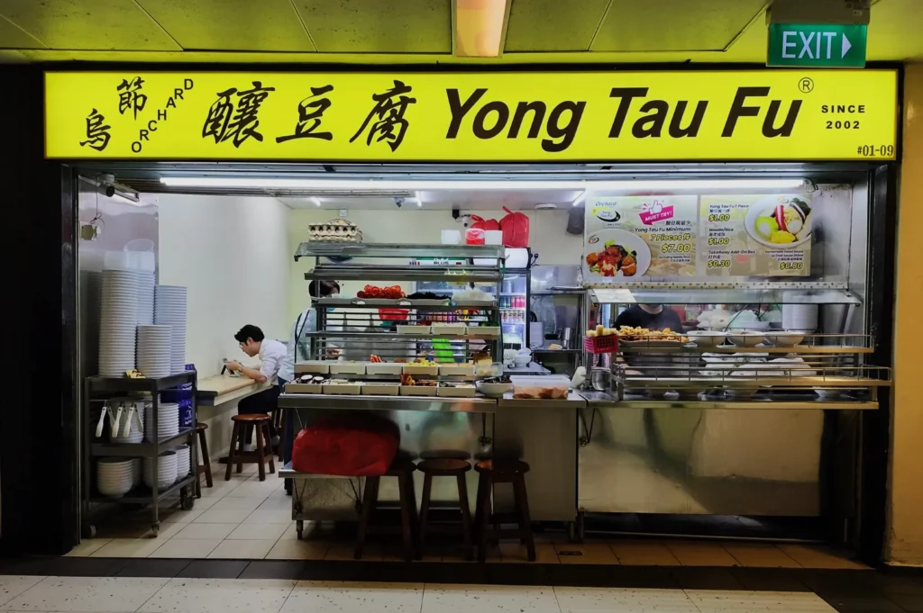 Street food stall with a bright yellow sign reading "Yong Tau Fu." Inside, a person seated on a stool, with dishes and ingredients on display. Cozy and inviting atmosphere.