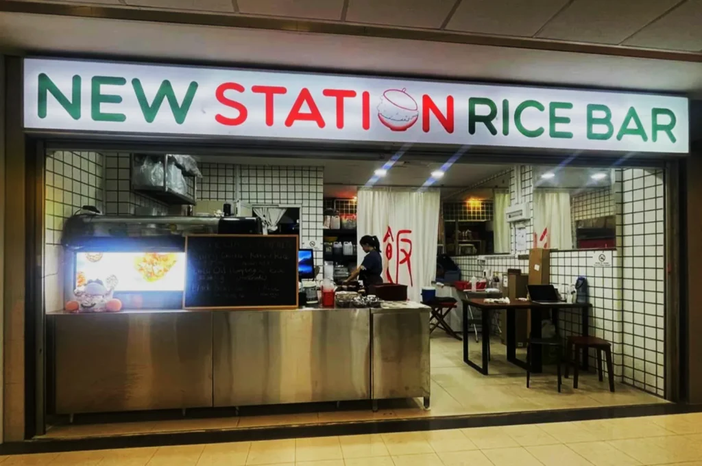 "Brightly lit storefront of 'New Station Rice Bar' with a worker behind a stainless steel counter, creating a welcoming and busy atmosphere."