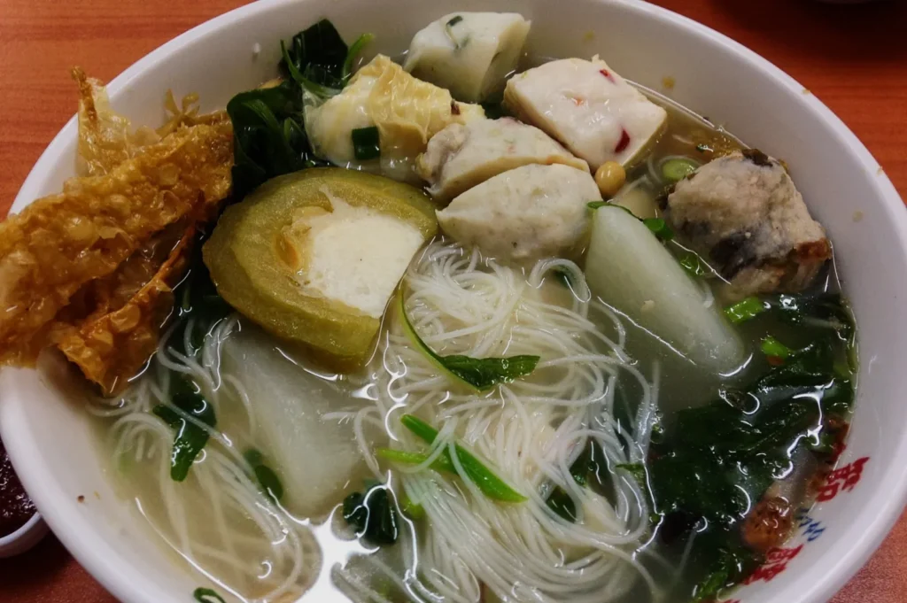 A steaming bowl of noodle soup with white vermicelli, assorted fish balls, leafy greens, fried tofu skin, and sliced vegetables in clear broth.
