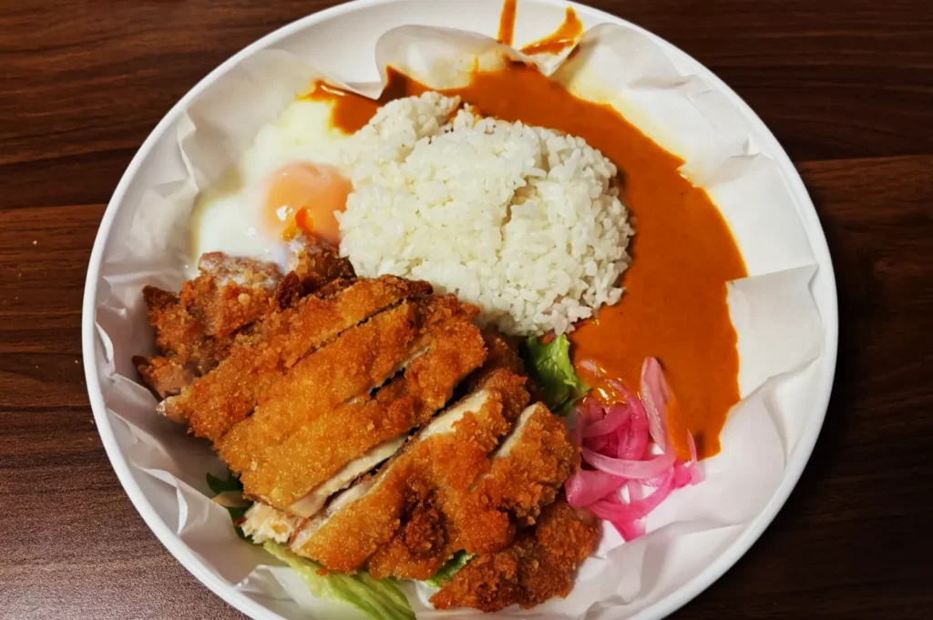 A plate of crispy fried chicken katsu, white rice topped with a runny poached egg, rich curry sauce, and pickled red onions. The dish looks appetizing and comforting.
