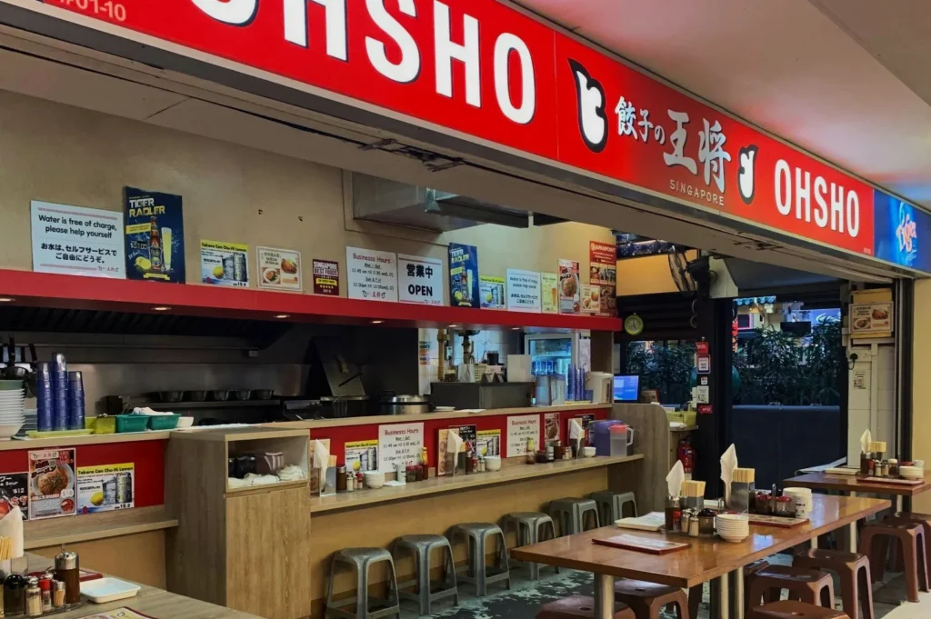Traditional Japanese eatery with a red "OHSHO" sign. Counter seating, simple decor, and outdoor table create a casual, inviting atmosphere.