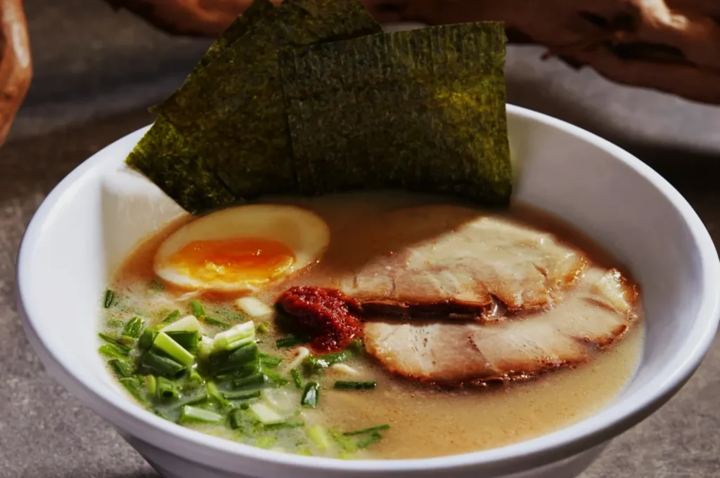 A bowl of ramen with rich broth, topped with two slices of chashu pork, a half-boiled egg, seaweed sheets, chopped green onions, and a dollop of red paste.