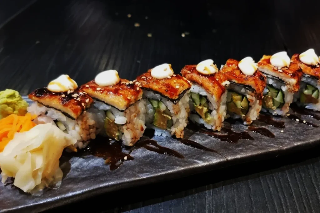 Sushi rolls with grilled eel and cream drizzle are artfully arranged on a black plate, accompanied by wasabi, pickled ginger, and shredded carrot.