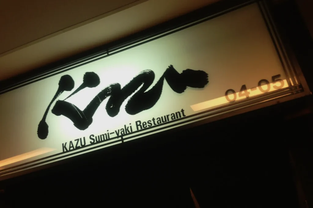 Illuminated sign for Kazu Sumi-yaki Restaurant with bold, artistic black lettering on a white background, evoking a modern and inviting ambiance.