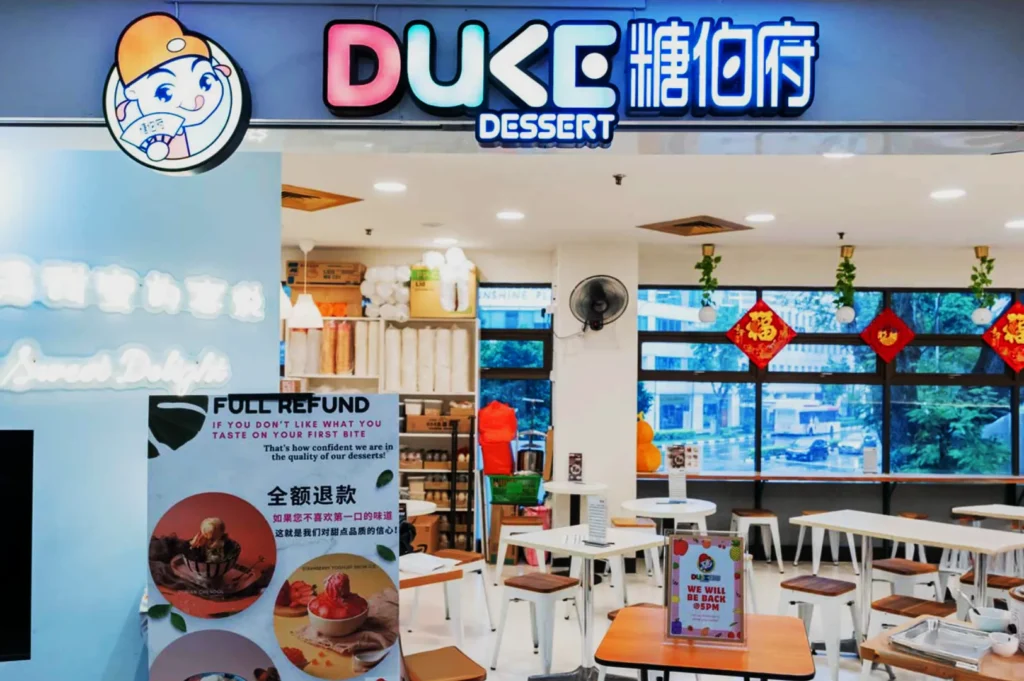 Brightly lit dessert shop interior with "DUKE DESSERT" sign. Tables, chairs, festive decorations, and a bold refund sign create a welcoming atmosphere.