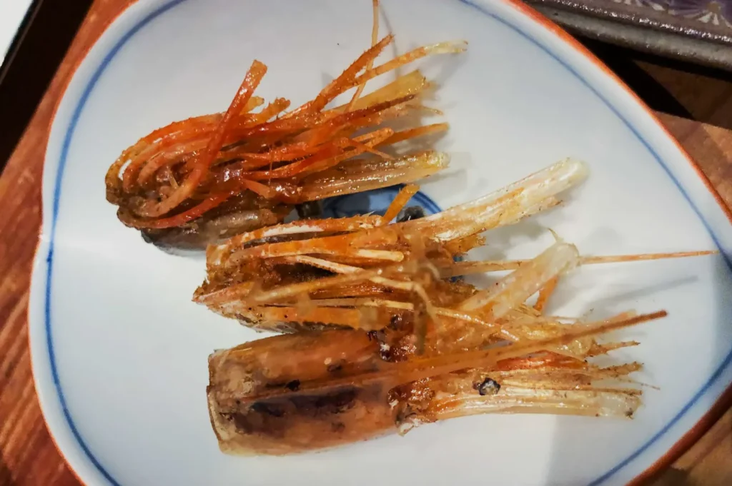 Fried shrimp heads on a white plate with blue rim, showcasing crispy orange and translucent textures. The dish appears delicate and crunchy.