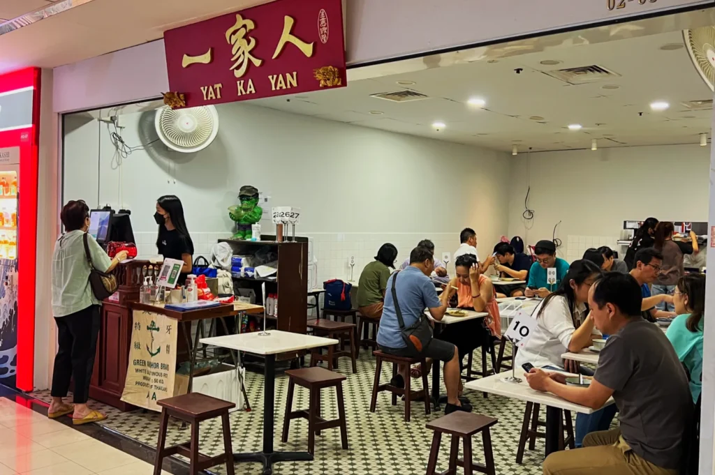 Bustling cafe with wooden tables and patrons seated, enjoying meals. A customer at the counter interacts with a cashier. Warm, inviting atmosphere.