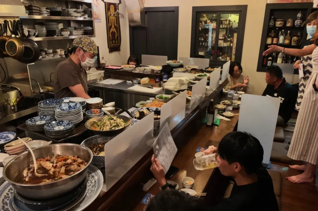 Chef in a cozy Japanese restaurant preparing traditional dishes. Diners seated at the bar enjoy their meals, chatting. Warm, inviting atmosphere.
