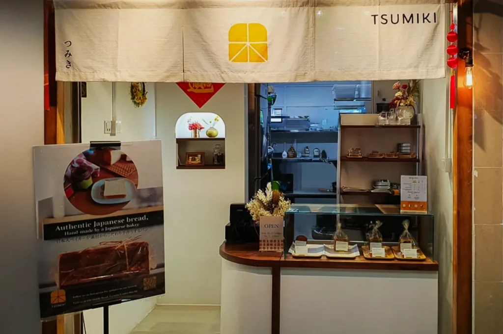 Alt text: "Small Japanese bakery Tsumiki with a stylish signboard and minimalistic decor. Display case with pastries and an 'Authentic Japanese bread' poster."