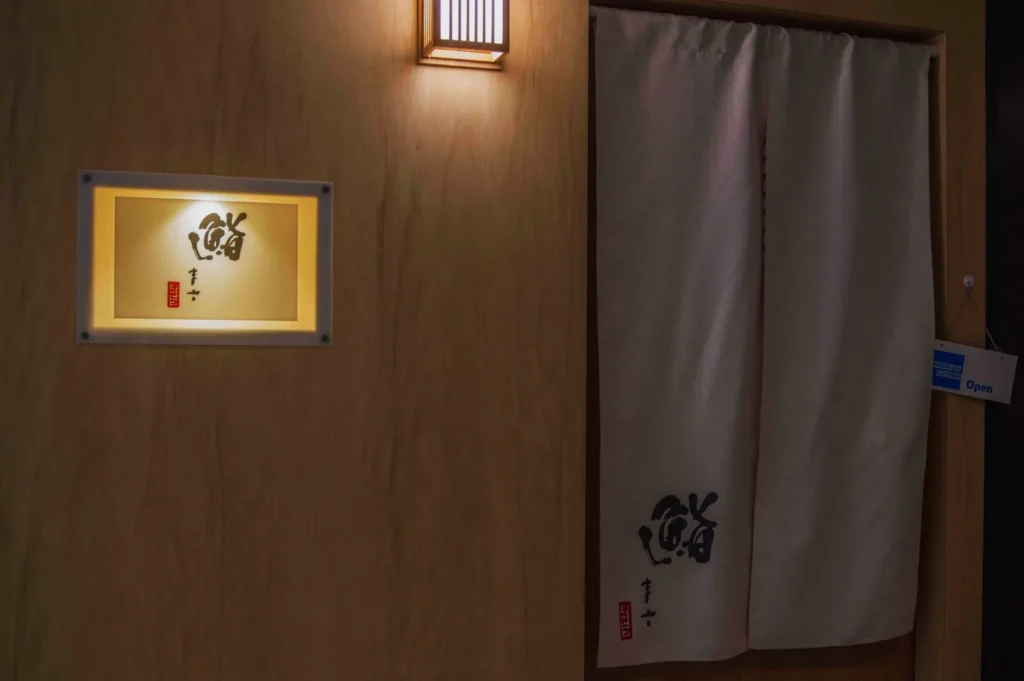 Dimly lit hallway with a beige curtain featuring Japanese calligraphy, a wooden wall with a lit sign displaying similar characters, and a simple lamp.