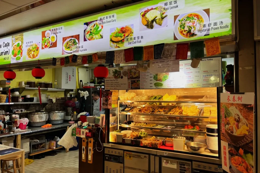 A vibrant vegetarian food stall in a market with colorful hanging lanterns and a menu board displaying dishes like Nasi Lemak and Thai Green Curry.