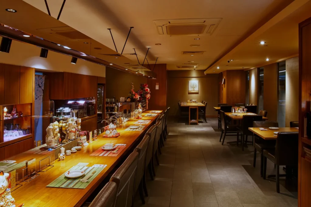 Warmly lit Japanese-style restaurant interior, featuring a long wooden counter adorned with decorative items, and neatly set tables in the background. Cozy and inviting ambiance.