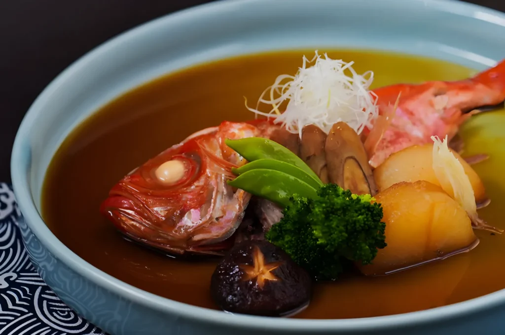 A colorful dish in a blue bowl features a cooked fish head garnished with slices of mushroom, broccoli, sugar snap peas, and potato atop a sauce.