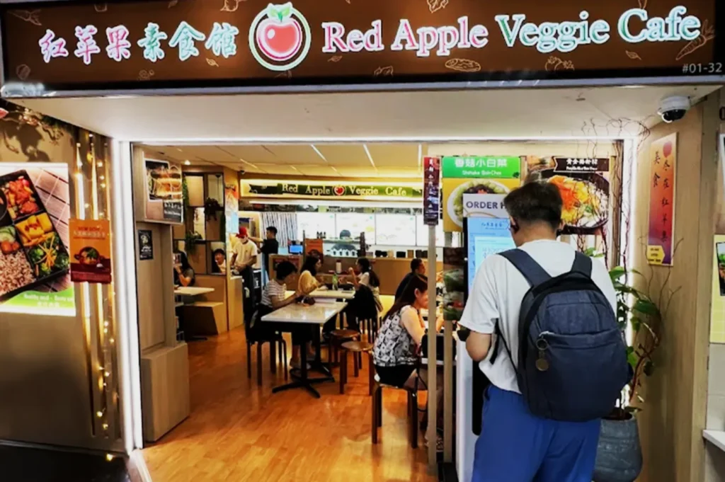 Entrance of Red Apple Veggie Cafe with a warm, inviting ambiance. Diners are seated at tables, and a person is ordering at a kiosk.