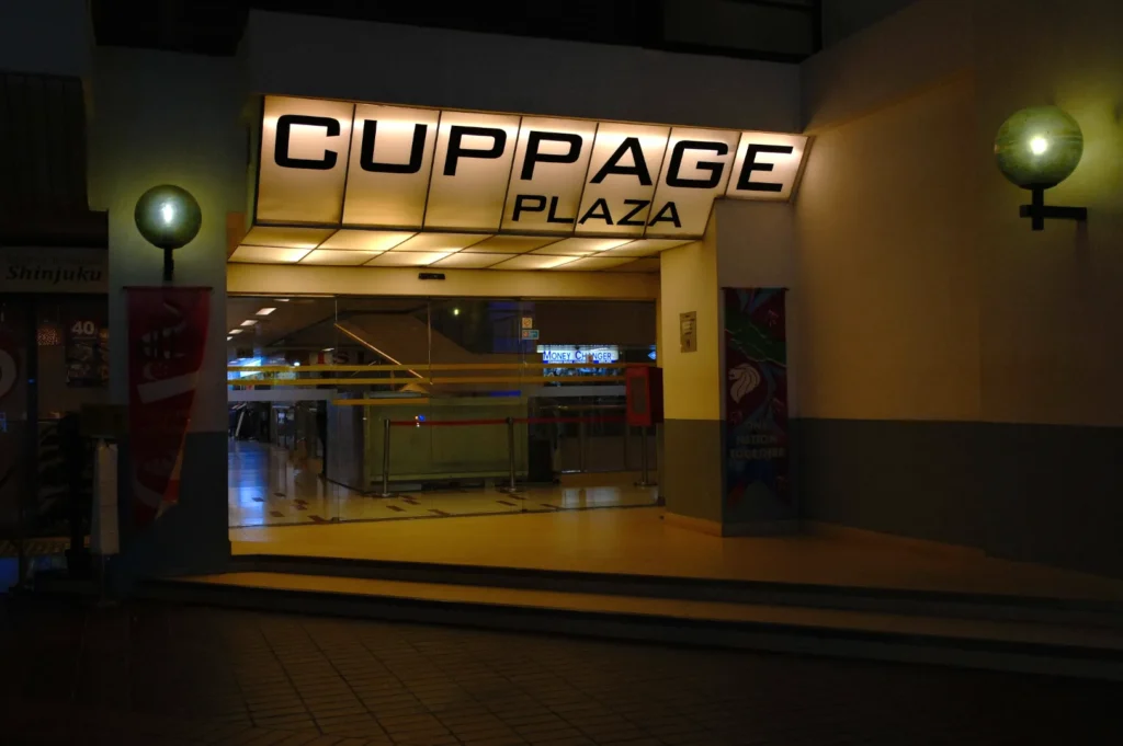 Entrance to Cuppage Plaza, warmly lit with glowing signage against the night sky. Modern glass doors lead into the building, flanked by spherical lights.