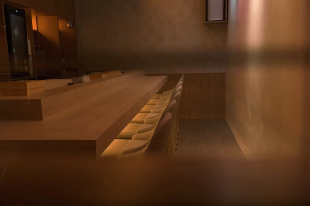 Warmly lit, minimalist room featuring a wooden bar counter with soft lighting underneath, lined with beige cushioned stools, creating an intimate ambiance.