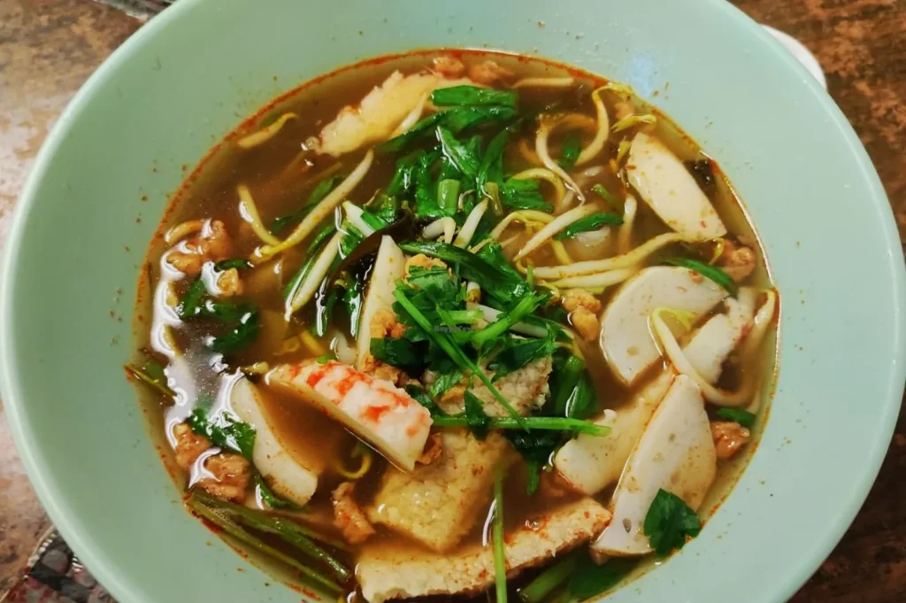 A steaming bowl of Vietnamese noodle soup with sliced fish cakes, crab sticks, bean sprouts, and fresh herbs, creating a warm, inviting meal.