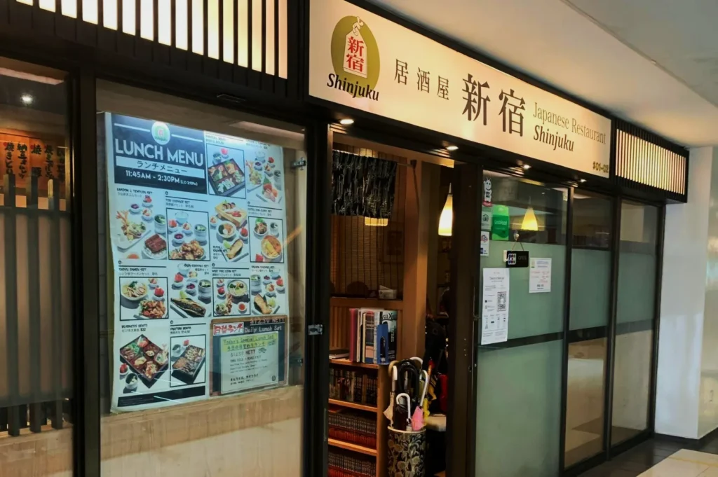 Street view of the entrance to Shinjuku Japanese Restaurant. A lunch menu poster with colorful dishes is displayed next to the glass door. Cozy ambiance.