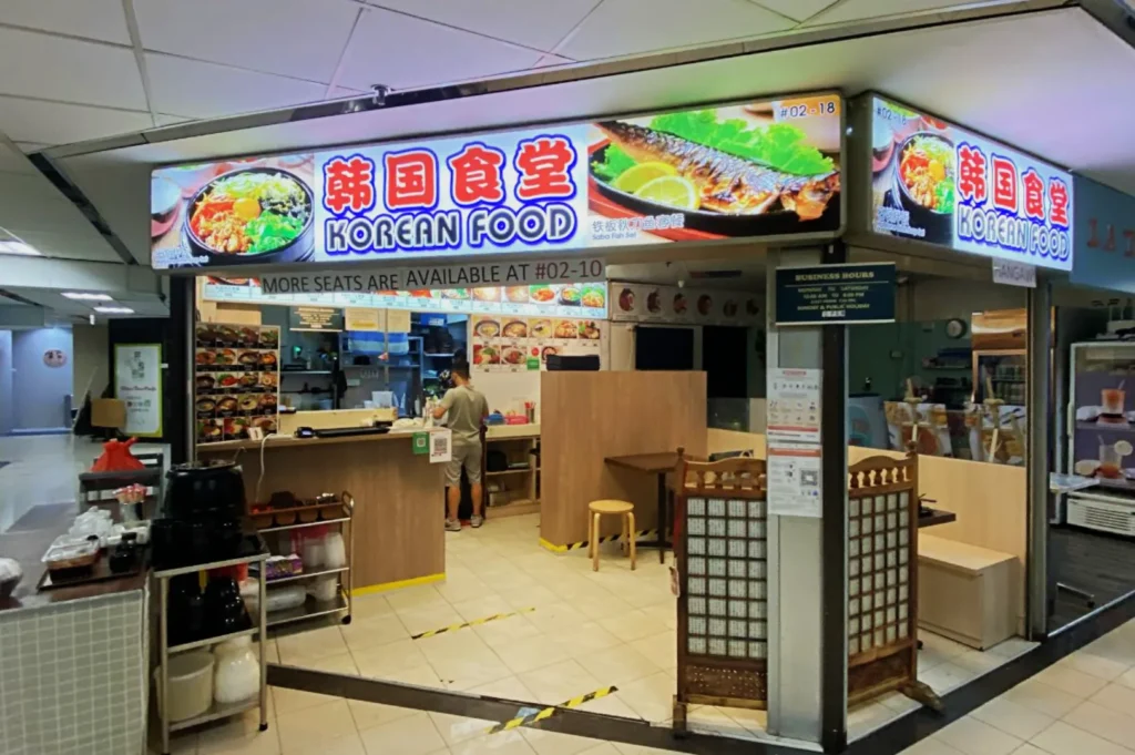 A small Korean food stall has a bright sign with images of dishes. Inside, a person works behind a counter surrounded by colorful menu pictures. Cozy and inviting.