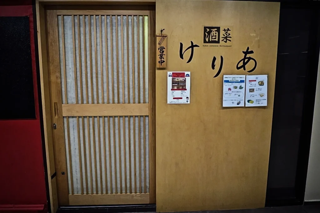 Wooden door with vertical slats next to a wall displaying Japanese text and restaurant posters. A small sign hangs, signaling the establishment is open.
