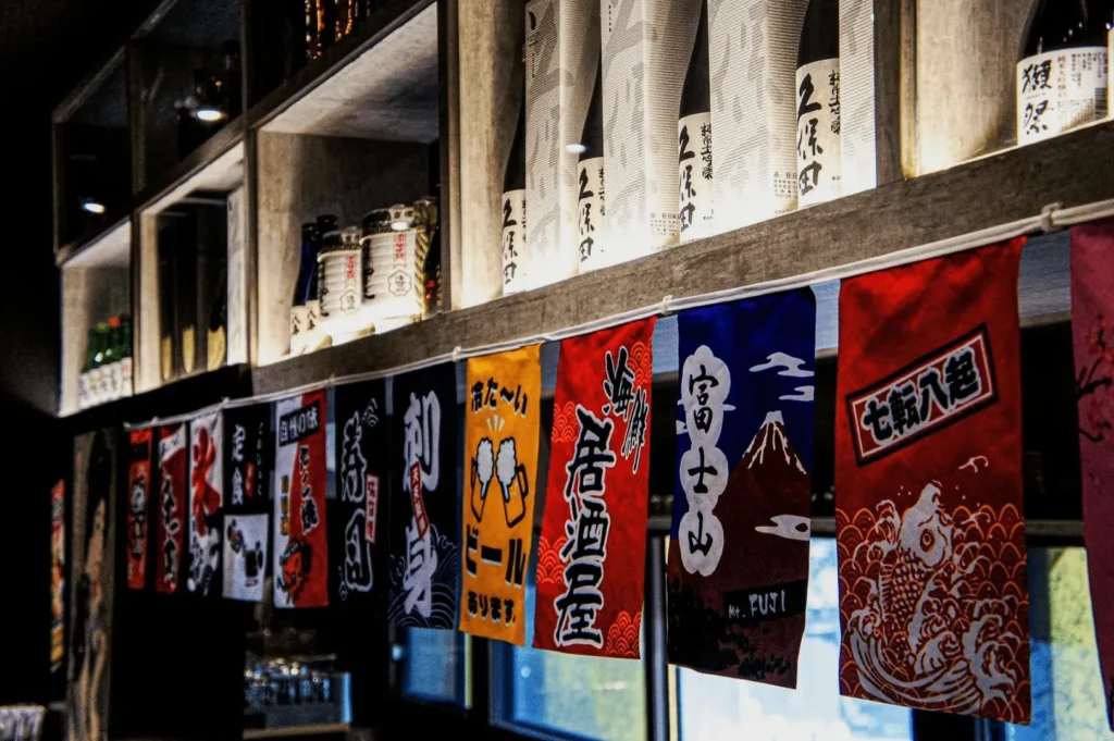 Colorful Japanese banners with bold text hang in a row beneath shelves stocked with bottles, creating a vibrant and traditional ambiance.
