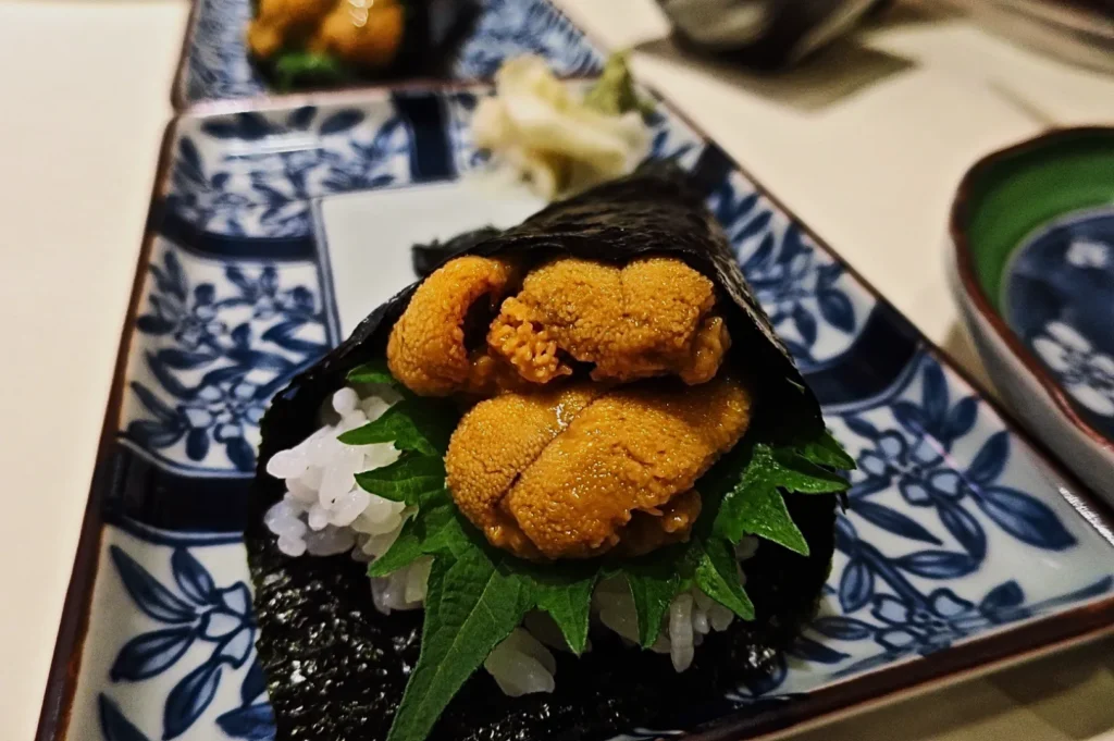 Close-up of a sushi hand roll with sea urchin, white rice, and shiso leaf wrapped in nori. Served on a blue-patterned plate with a calm dining ambiance.