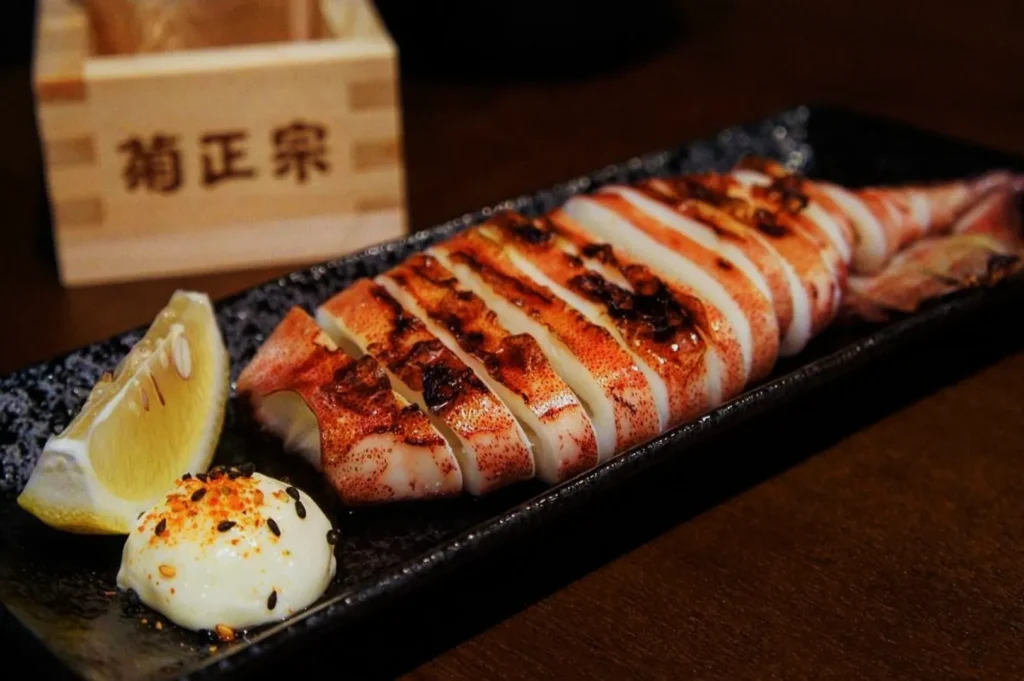 Grilled squid sliced and arranged on a black rectangular plate with a lemon wedge and seasoned mayonnaise. A wooden box with Japanese writing is in the background.