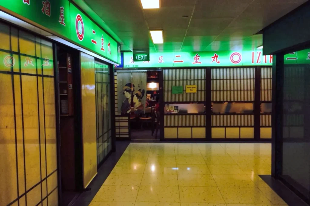 Corridor leading to a Japanese restaurant with a green neon sign. The entrance has traditional decor and an illuminated exit sign above. Cozy ambiance.